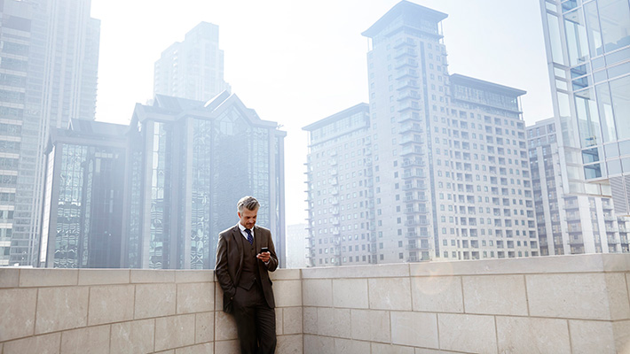 Businessman in a suit checking phone on a rooftop terrace, surrounded by urban skyline of tall modern buildings 