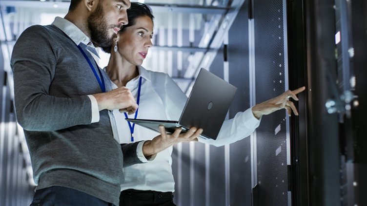 Two people focused on a laptop in a server room, surrounded by rows of servers and technical equipment.