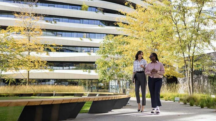 Two people walk leisurely in a park, surrounded by greenery, with a building standing in the background.