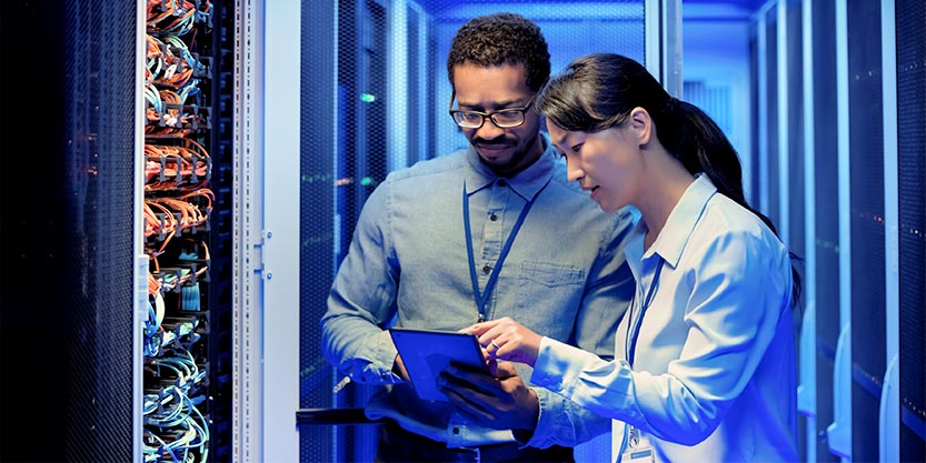 Two individuals examining a computer setup within a server room filled with equipment and servers.