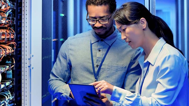 Two individuals examining a computer setup within a server room filled with equipment and servers.