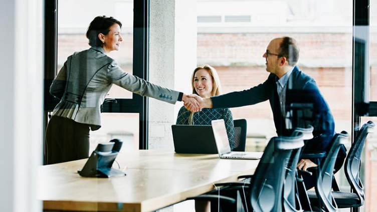 Two professionals shaking hands across a conference room table with another professional smiling