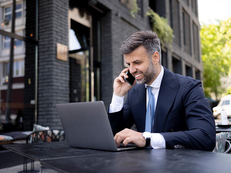 A professional is talking on a mobile phone while working on a laptop.