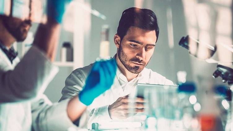 Two men wearing lab coats studying a tablet together, demonstrating collaboration in a scientific environment.