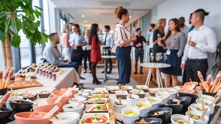 A group of colleagues socializing with food and drink in the office at a workplace event.