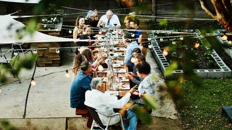 A diverse group of people of mixed ages sitting around a table  together in a communal garden space.